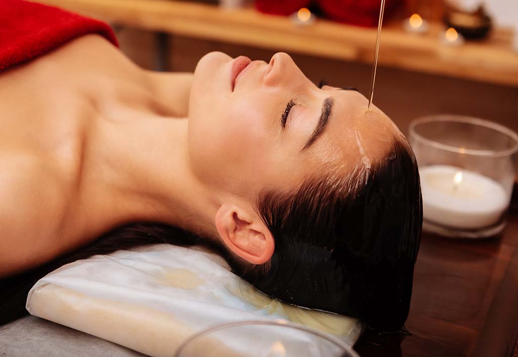 Feeling meditative. Flawless good-looking woman having thin stream of oil pouring on her face while she having self-care routine